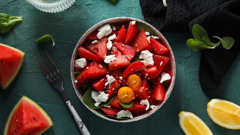 Erfrischende Sommerrezepte: Schüssel mit Wassermelone und Feta. Steht auf grünem Hintergrund. Es liegen Wassermelonenstücke und Zitronenstücke daneben.
