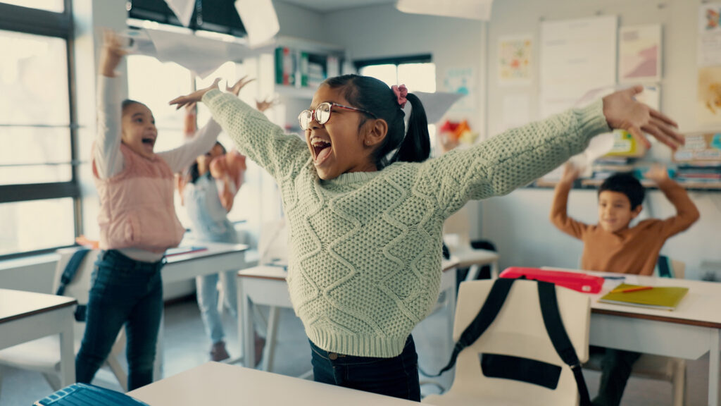 Semesterferien: Begeisterte Grundschulkinder bewegen sich aktiv im Klassenraum und werfen Papier in die Luft.