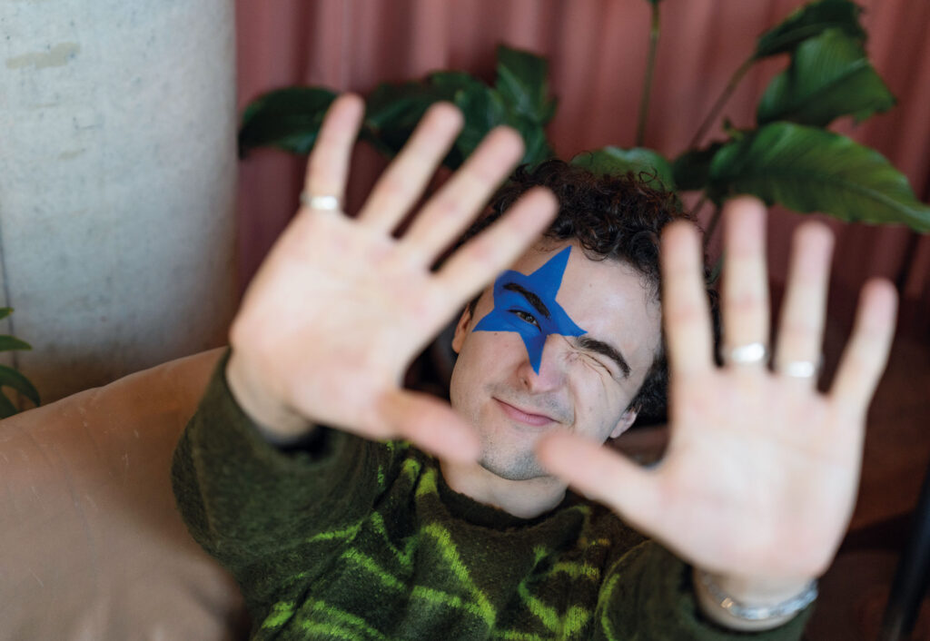 Portrait von COSMÓ mit ausgestreckten Händen in Richtung Kamera und zugezwinkertem Auge.