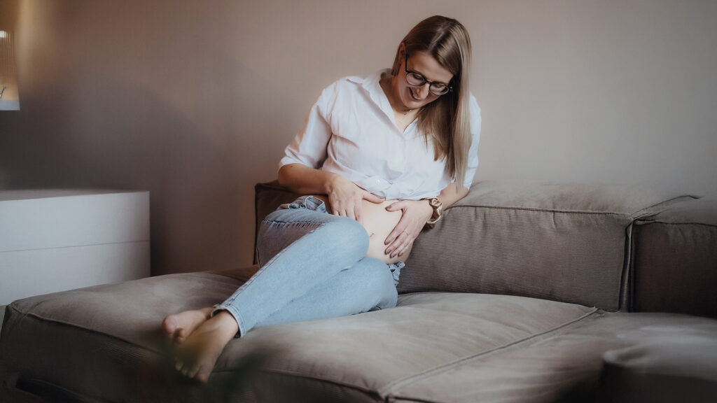 Schwanger: Eine schwangere Frau sitzt auf einem Sofa und legt die Hände auf ihren Bauch.