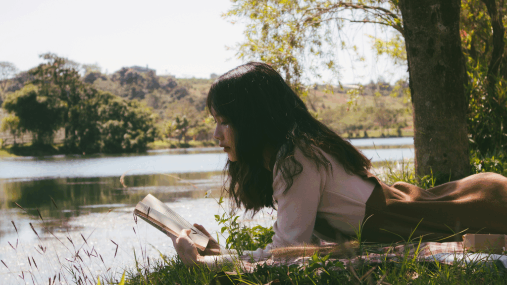 Eine Frau liegt auf dem Bauch und liest ein Buch. Im Hintergrund sind man einen See und Bäume